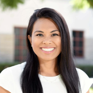 A woman with long black hair wearing a white blouse standing in front of a neutral background