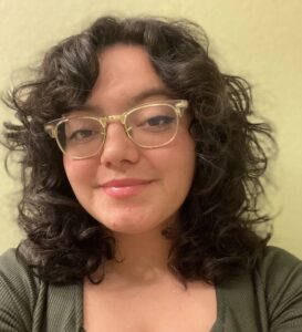 A femme presenting person with curly brown hair wearing glasses and a green dress against a neutral background