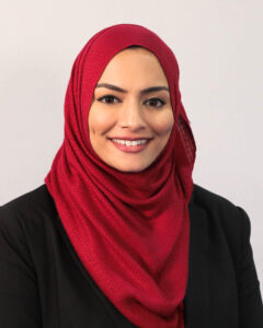 A woman wearing a red hijab and a black blouse standing in front of a neutral background