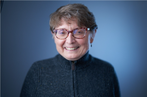 A woman with short light brown hair with multi-colored glasses smiling wearing a blue zip up against a neutral background.