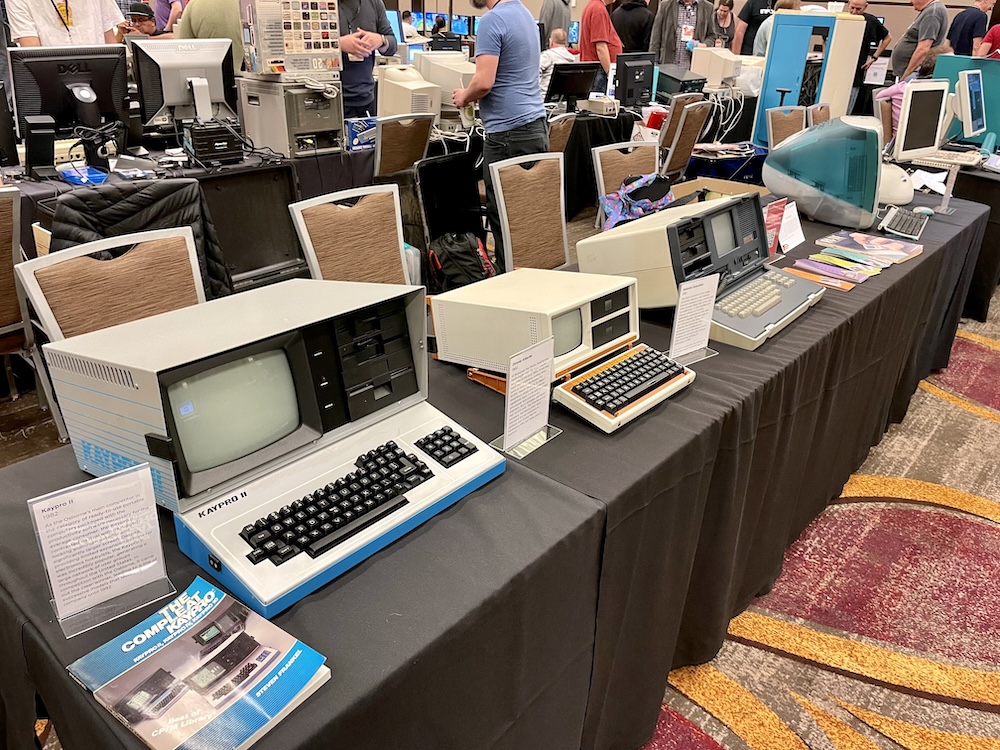 Photo of an exhibition table containing three luggable computers and two early iMacs.
