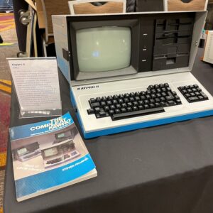 A large luggable computer in a metal case in open position sitting next to a manual for that model. The model is the Kaypro II. The text of the sign next to it is in the caption below.