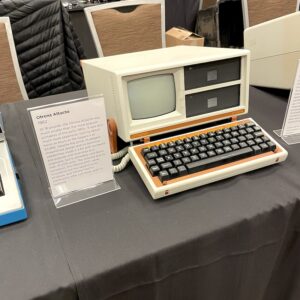 A small orange and white luggable computer resting on the table in open position. The model is the Otrona Attaché. The text of the sign next to it is in the caption below.