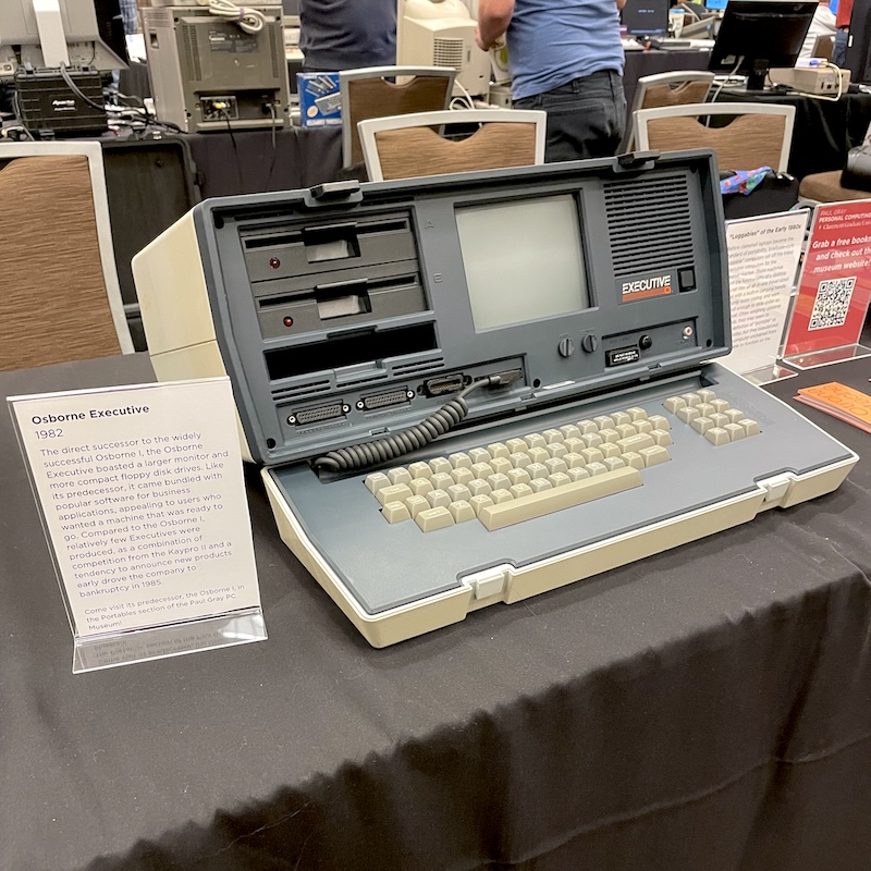 A large suitcase-shaped luggable computer, resting on the table in open position. The model is the Osborne Executive. The text of the sign next to it is in the caption below.