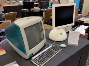 A Bondi Blue iMac G3 with keyboard and mouse and an iMac G4 next to a small sign. The text of the sign is in the caption below.